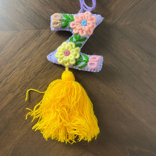 Handmade embroidered letter Z ornament with floral stitching on purple fabric and bright yellow tassel on wooden background