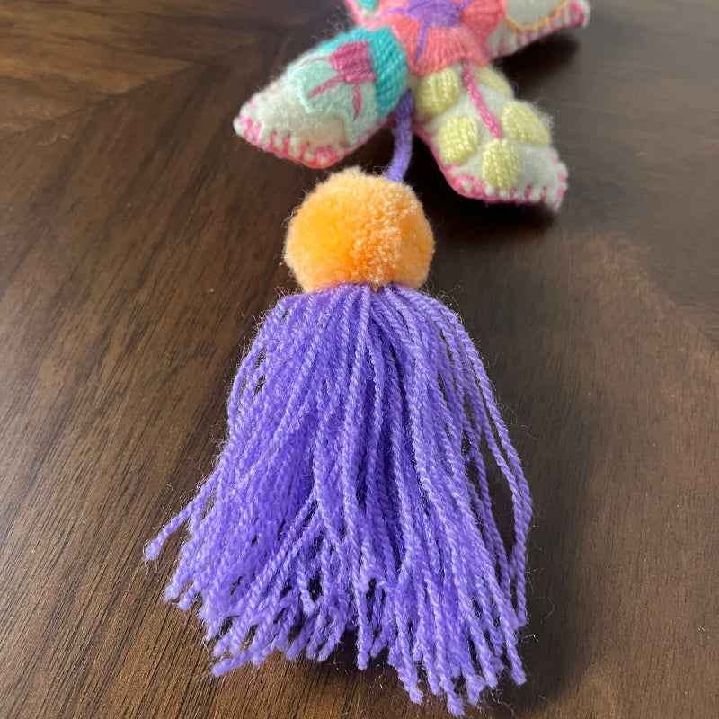 Colorful handmade embroidered letter X ornament with floral patterns, yellow pom pom and purple tassel on wooden background