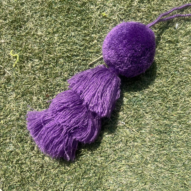Dark purple fluffy tassel keychain with soft pom pom placed on grass background