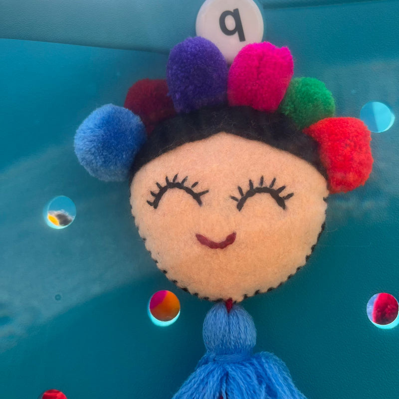 Handmade smiling Frida-inspired doll charm with colorful pom-poms and blue tassel on teal bag