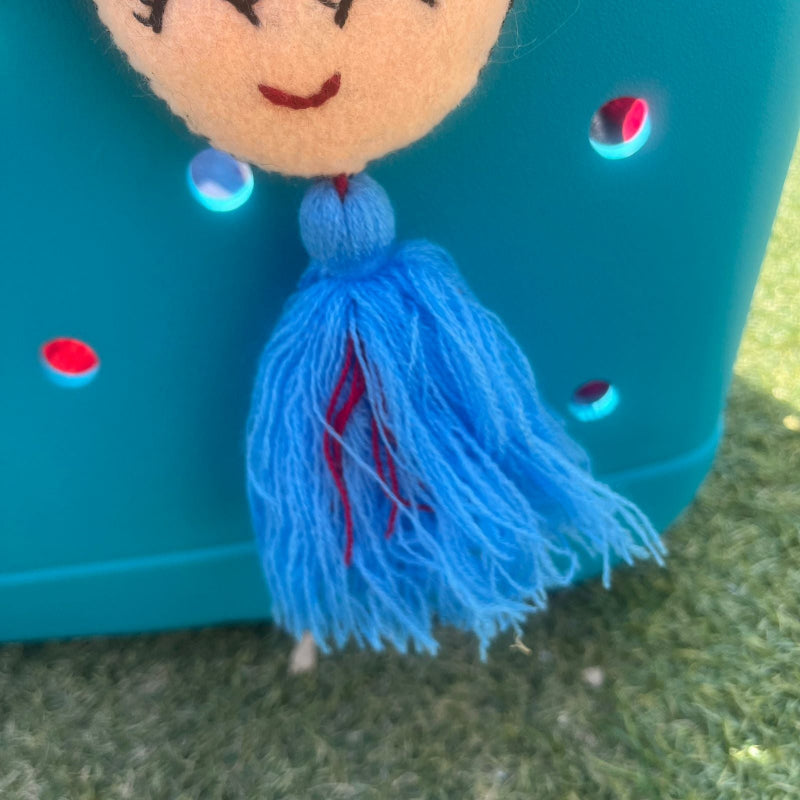 Handmade smiling Frida-inspired doll charm with colorful pom-poms and blue tassel on teal bag