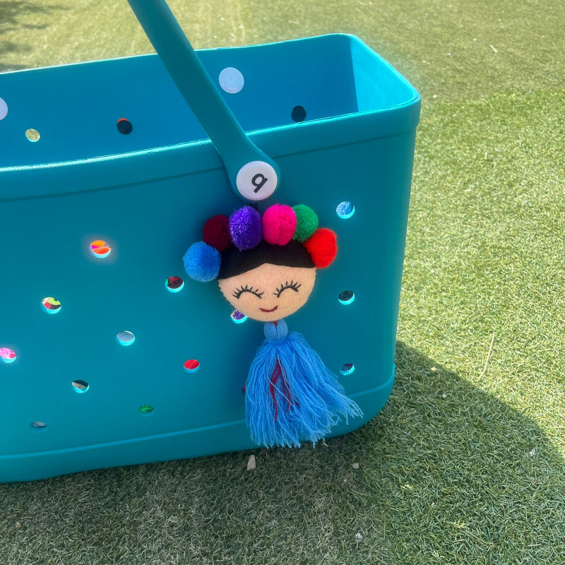 Handmade smiling Frida-inspired doll charm with colorful pom-poms and blue tassel on teal bag