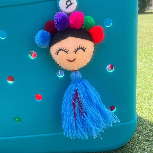Handmade smiling Frida-inspired doll charm with colorful pom-poms and blue tassel on teal bag