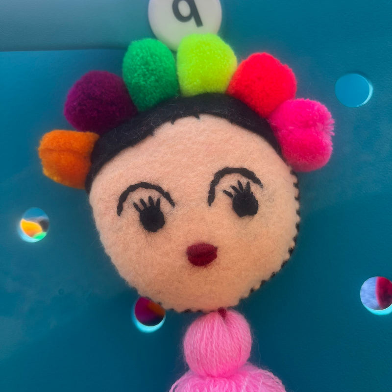 Handmade Frida-inspired doll charm with colorful pom-poms and soft pink tassel on teal bag