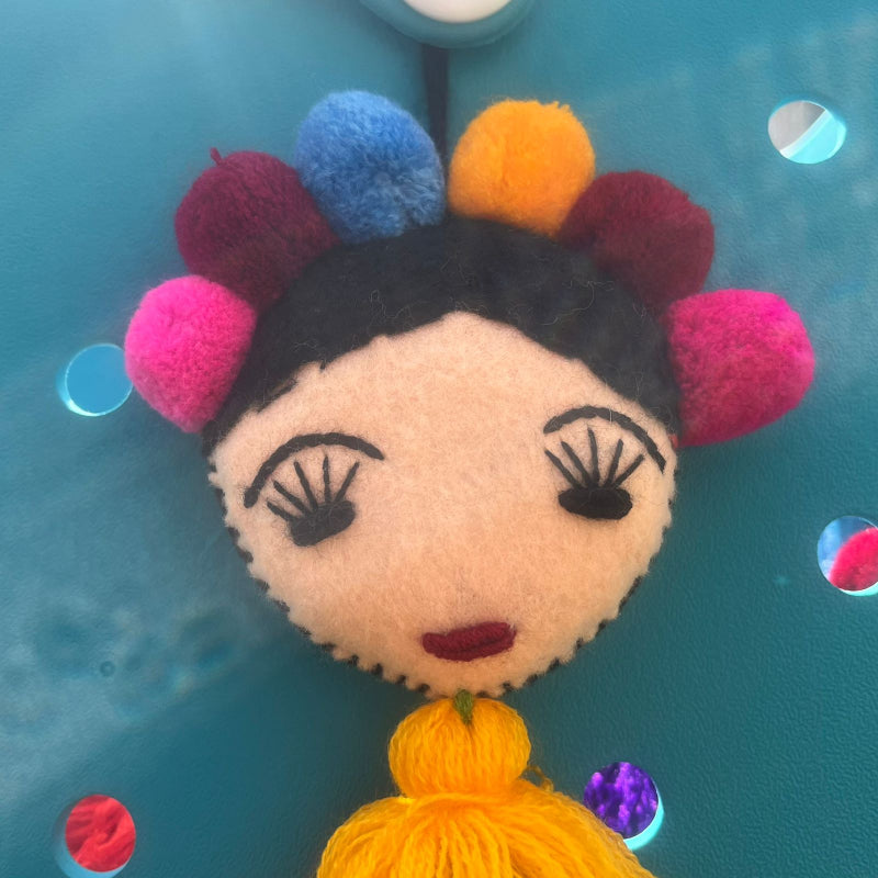 Handmade Frida-inspired doll charm with colorful pom-poms and yellow tassel on teal bag