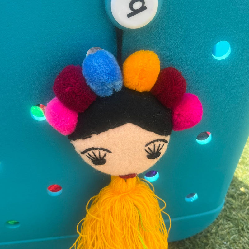 Handmade Frida-inspired doll charm with colorful pom-poms and yellow tassel on teal bag