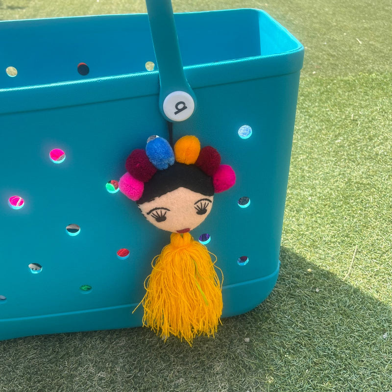 Handmade Frida-inspired doll charm with colorful pom-poms and yellow tassel on teal bag