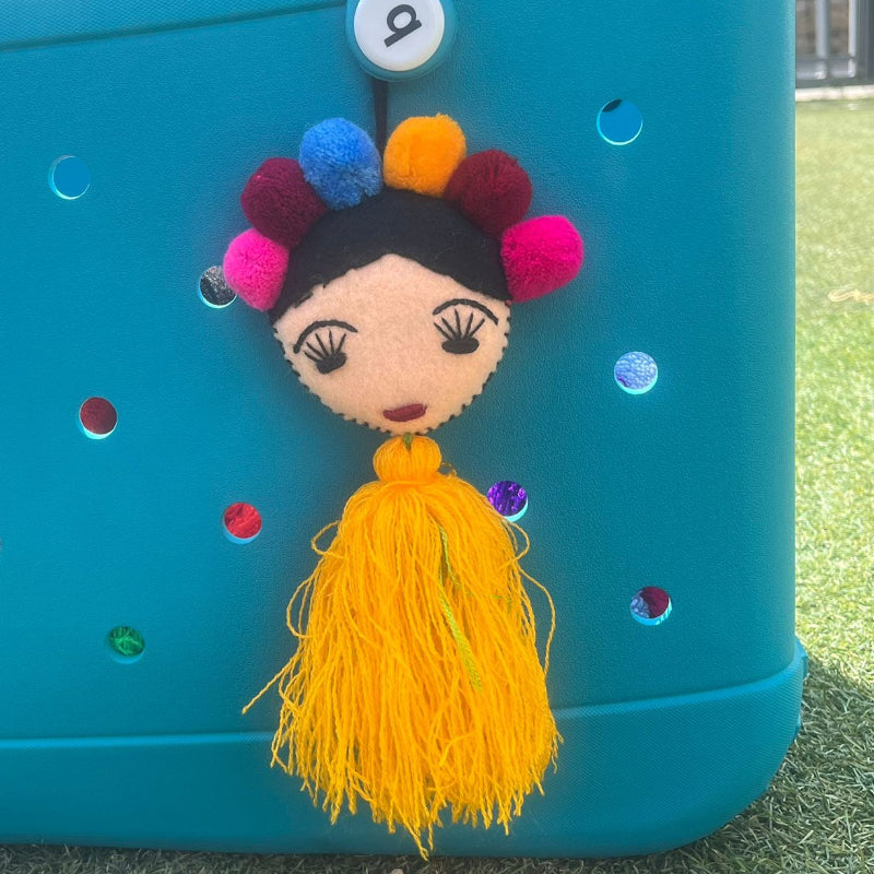 Handmade Frida-inspired doll charm with colorful pom-poms and yellow tassel on teal bag