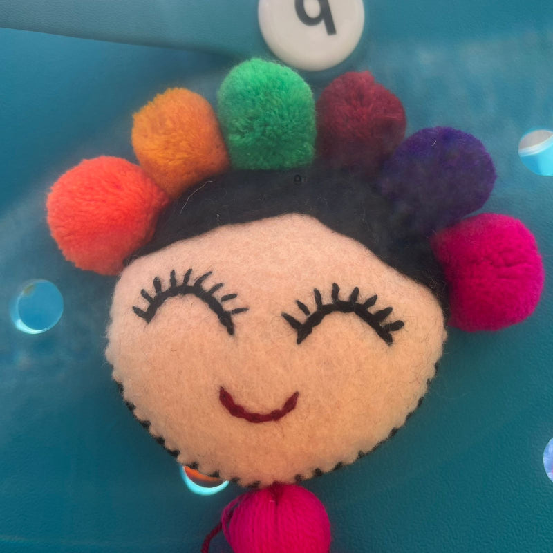 Handmade smiling Frida-inspired doll charm with colorful pom-poms and pink tassel on teal bag