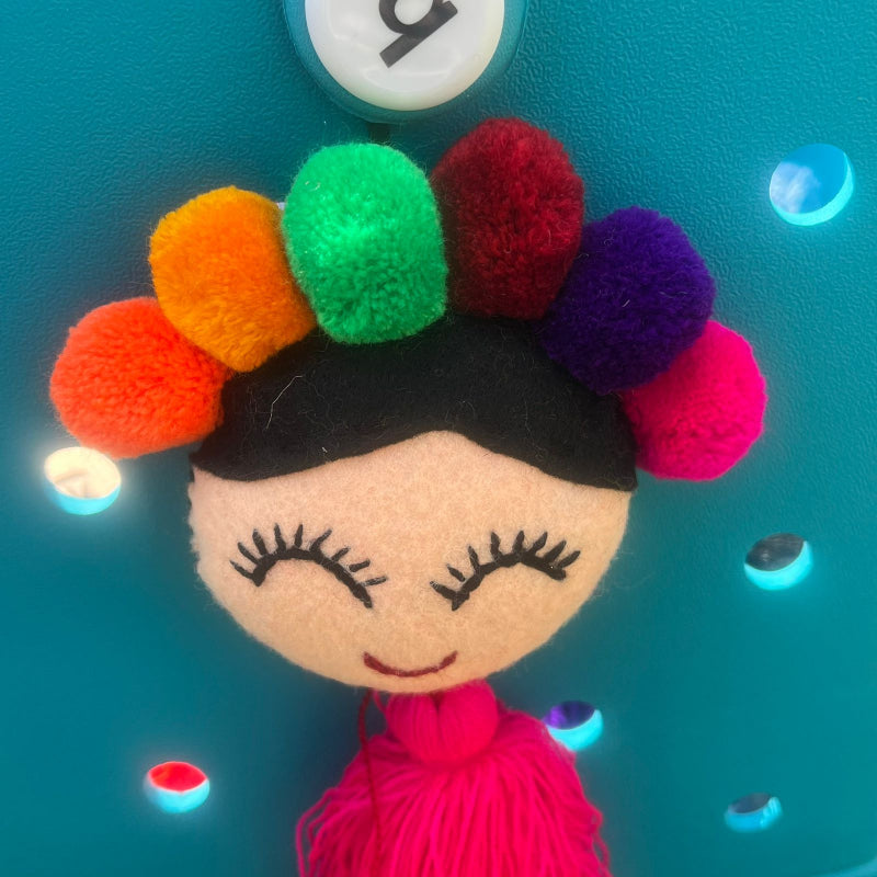 Handmade smiling Frida-inspired doll charm with colorful pom-poms and pink tassel on teal bag