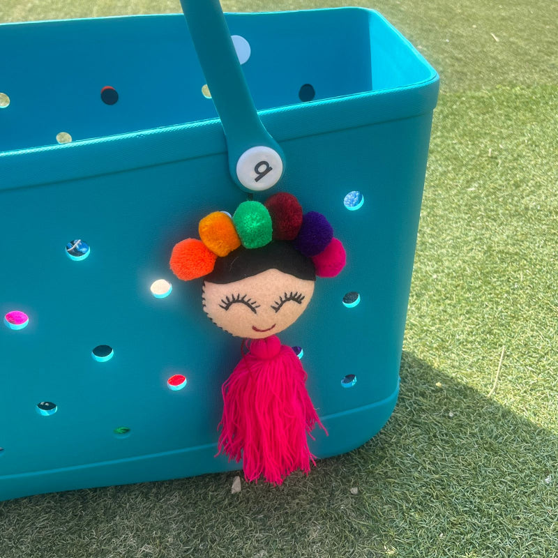 Handmade smiling Frida-inspired doll charm with colorful pom-poms and pink tassel on teal bag