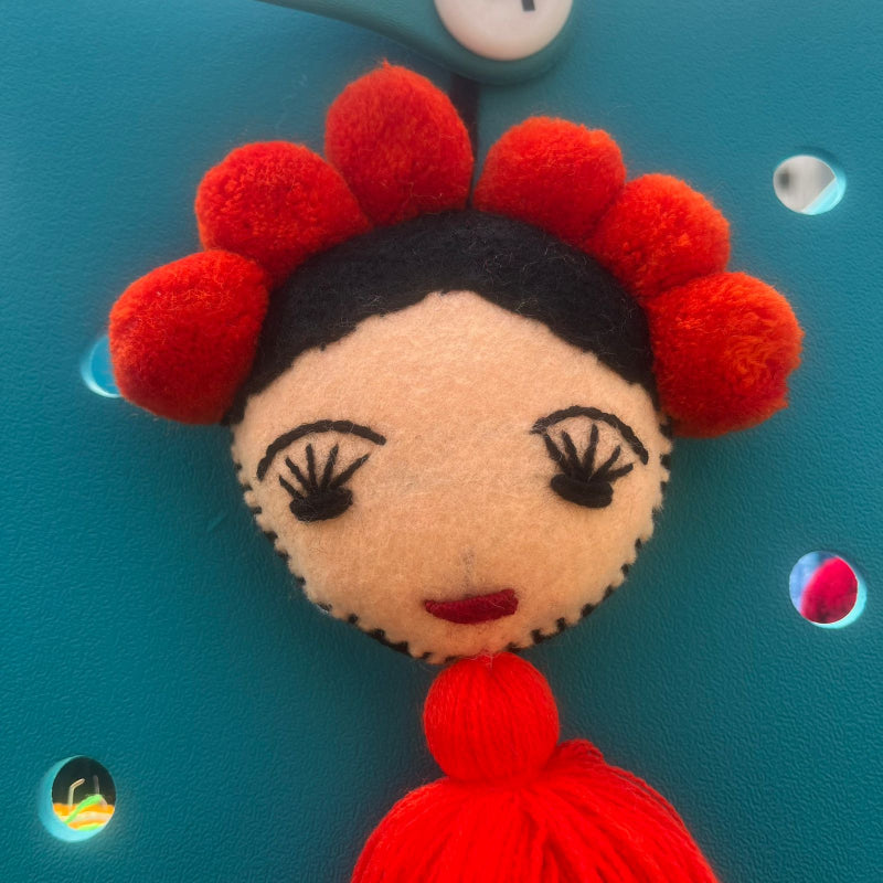 Handmade Frida-inspired doll charm with red pom-poms and red tassel on teal bag