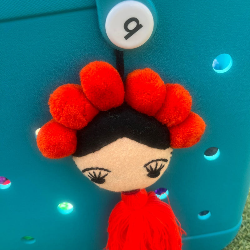 Handmade Frida-inspired doll charm with red pom-poms and red tassel on teal bag