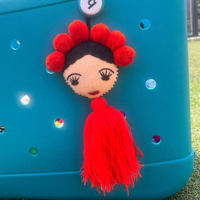 Handmade Frida-inspired doll charm with red pom-poms and red tassel on teal bag
