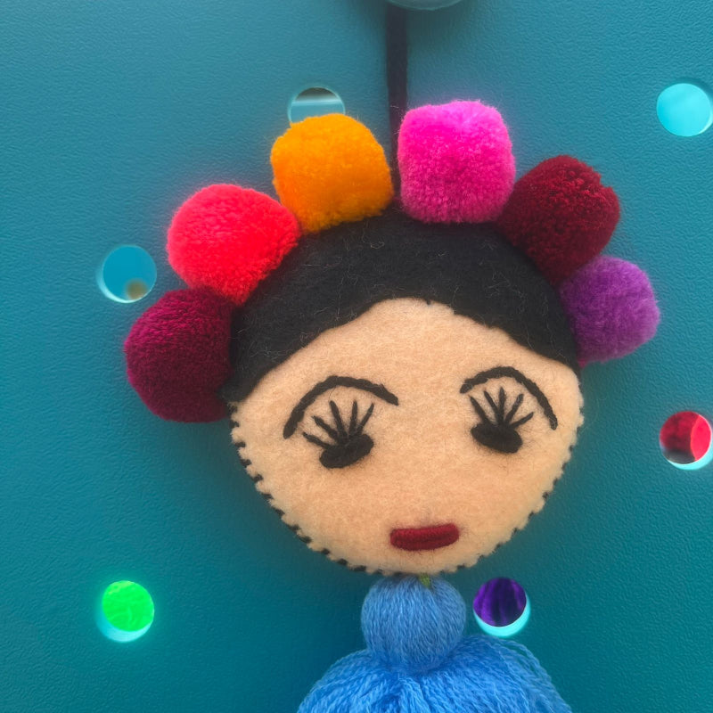 Handmade Frida-inspired doll ornament with colorful pom-poms and blue tassel attached to teal bag