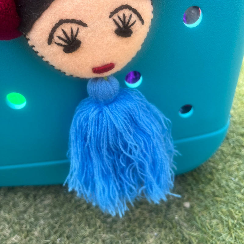 Handmade Frida-inspired doll ornament with colorful pom-poms and blue tassel attached to teal bag