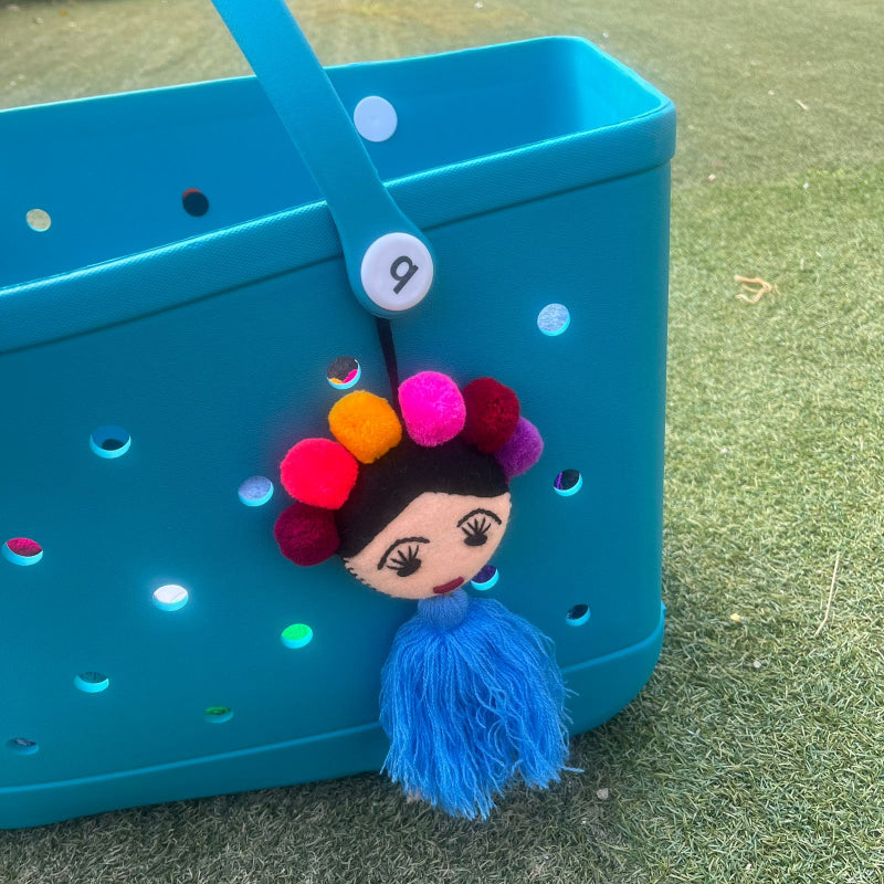 Handmade Frida-inspired doll ornament with colorful pom-poms and blue tassel attached to teal bag