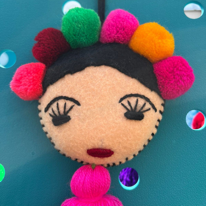 Handmade Frida-inspired doll charm with colorful pom-poms and pink tassel on teal bag