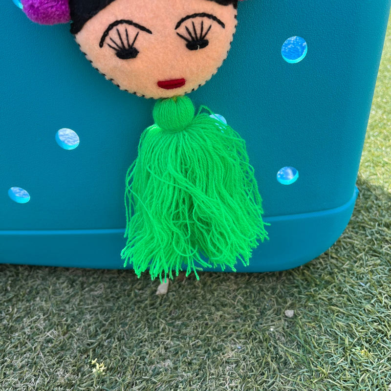 Handmade Frida-inspired doll charm with colorful pom-poms and green tassel attached to teal bag