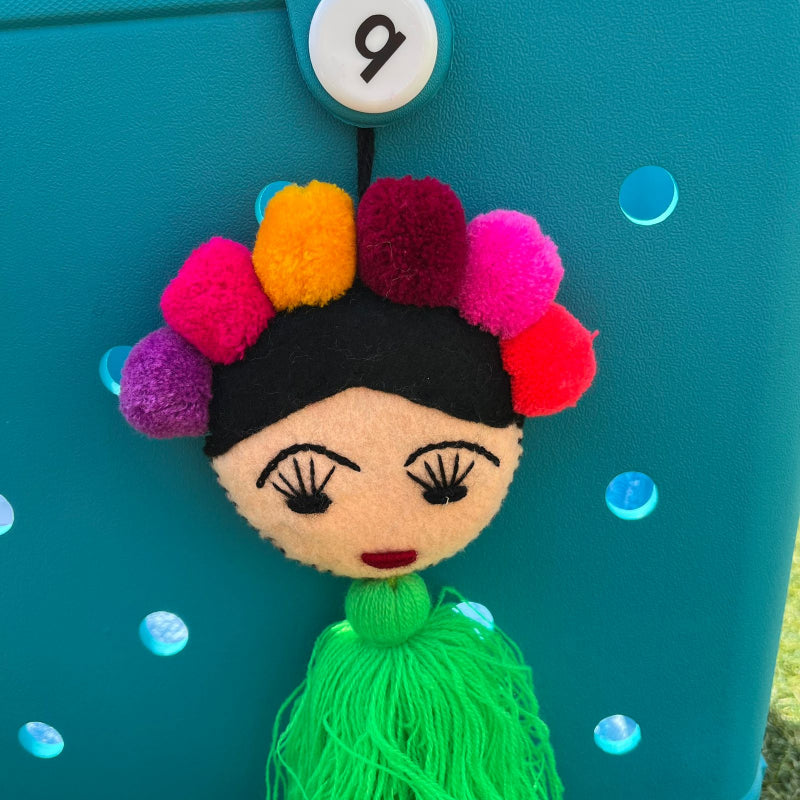 Handmade Frida-inspired doll charm with colorful pom-poms and green tassel attached to teal bag