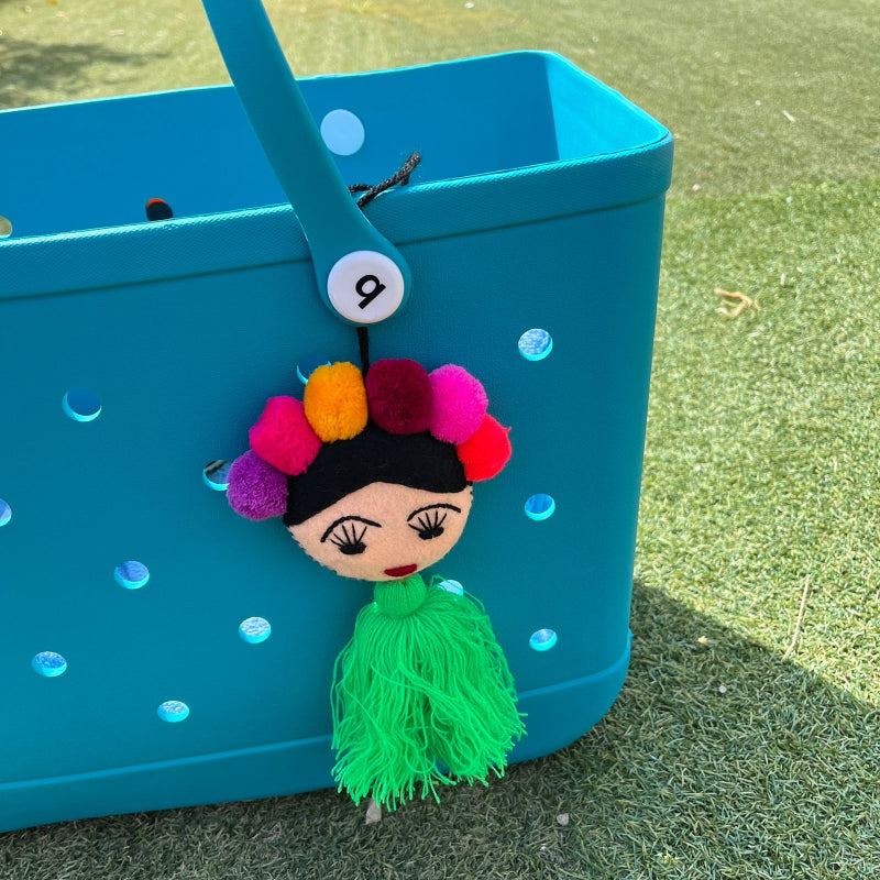 Handmade Frida-inspired doll charm with colorful pom-poms and green tassel attached to teal bag