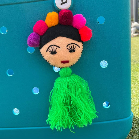 Handmade Frida-inspired doll charm with colorful pom-poms and green tassel attached to teal bag