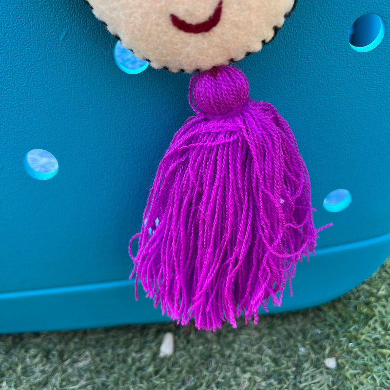 Handmade smiling Frida-inspired doll charm with colorful pom-poms and purple tassel on teal bag
