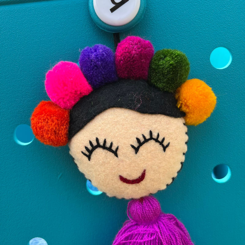 Handmade smiling Frida-inspired doll charm with colorful pom-poms and purple tassel on teal bag