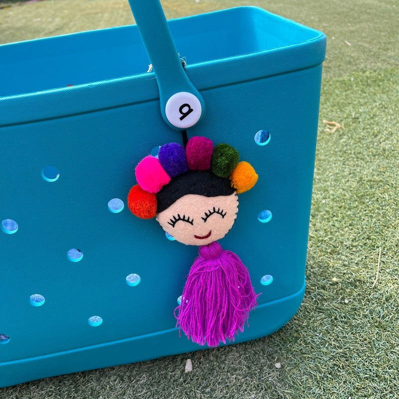 Handmade smiling Frida-inspired doll charm with colorful pom-poms and purple tassel on teal bag
