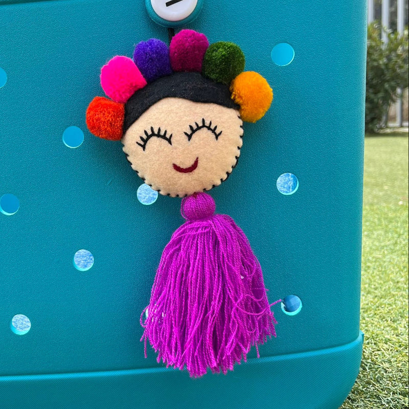 Handmade smiling Frida-inspired doll charm with colorful pom-poms and purple tassel on teal bag