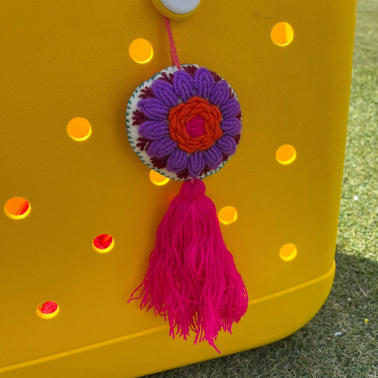 Handmade embroidered floral ornament with purple petals, orange center, and pink tassel on yellow background
