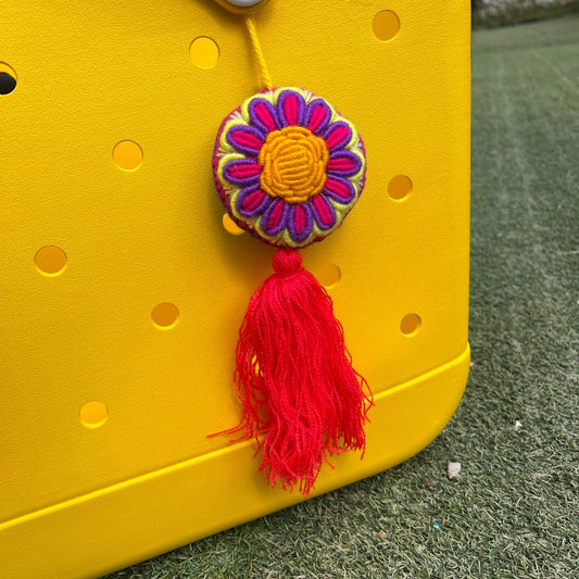 Handmade embroidered floral ornament with yellow center, pink and purple petals, and red tassel on a yellow background