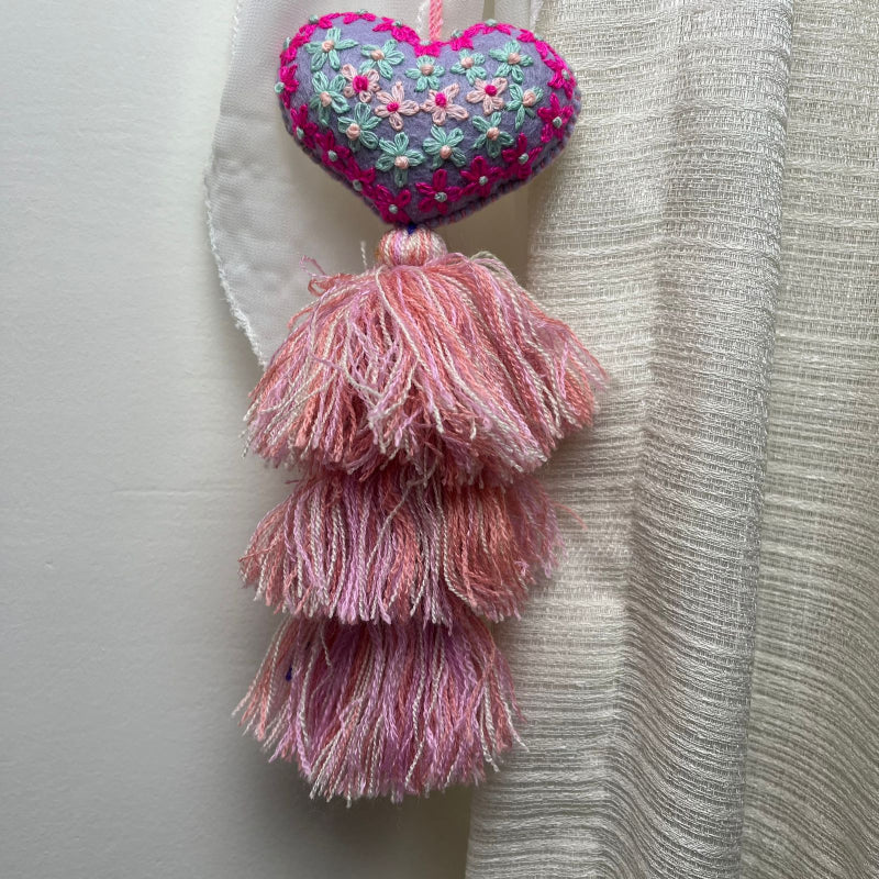 Handmade lavender embroidered heart tassel with colorful floral embroidery and pink yarn fringe on curtain