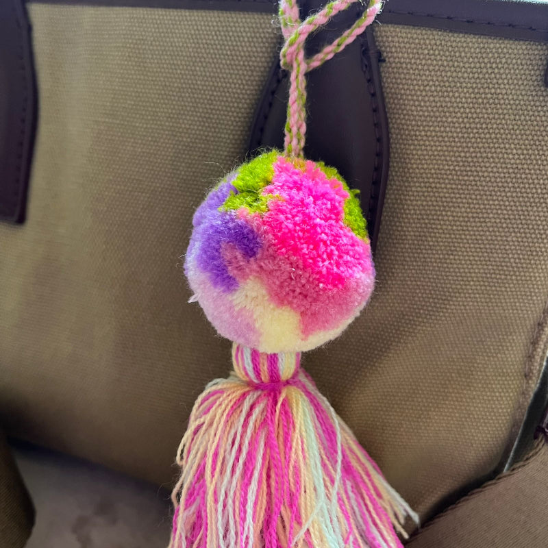 Soft pink pom pom tassel bag charm with layered pastel tassels attached to a beige handbag