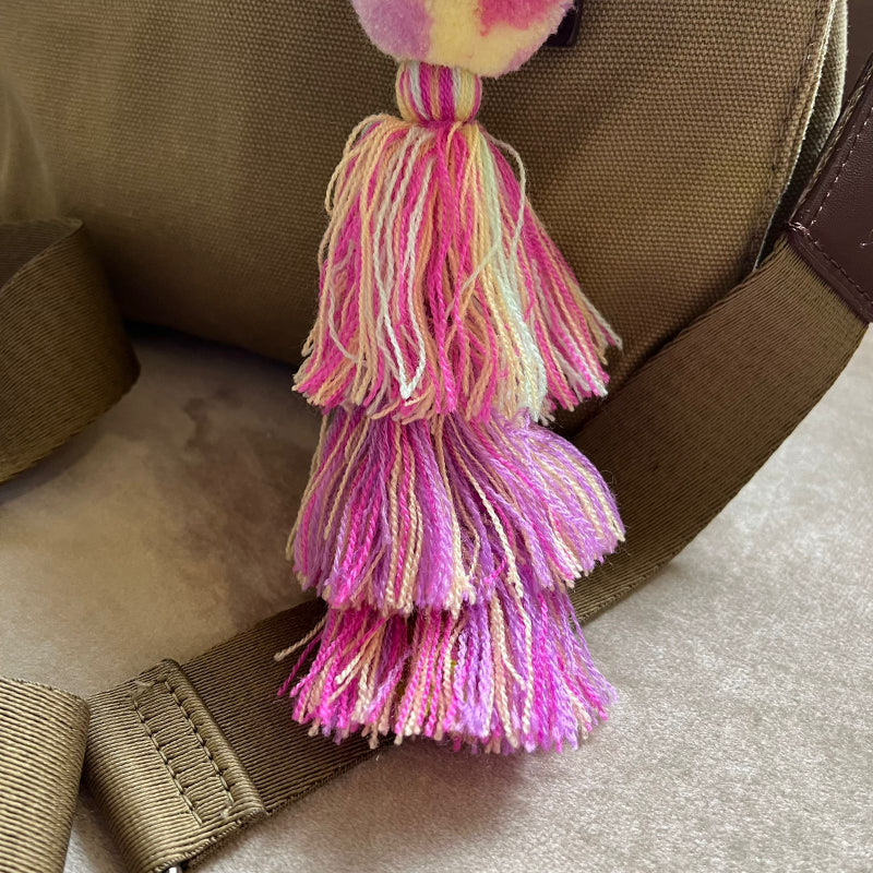 Soft pink pom pom tassel bag charm with layered pastel tassels attached to a beige handbag