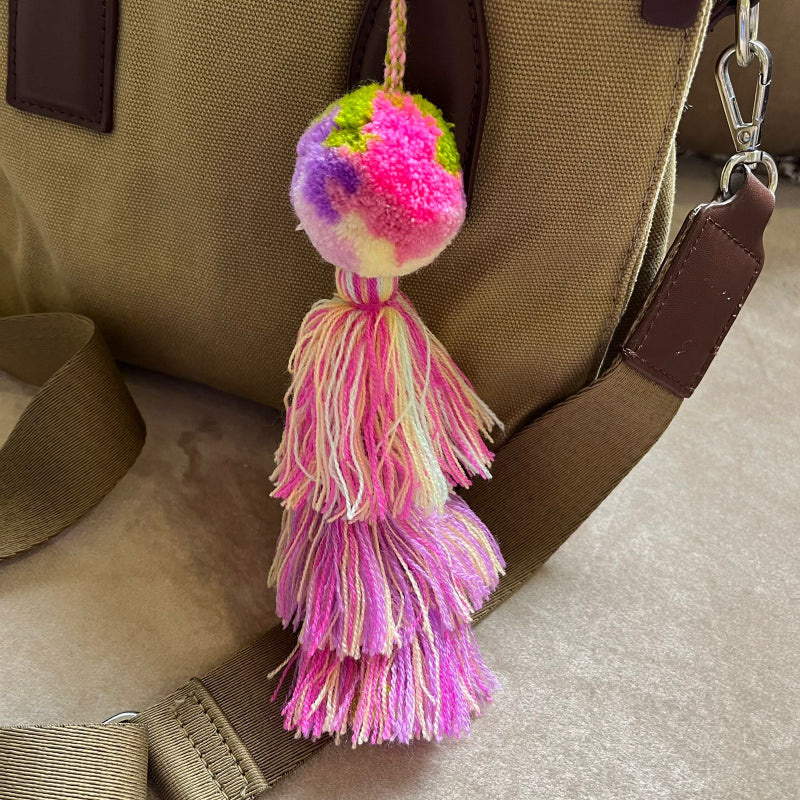 Soft pink pom pom tassel bag charm with layered pastel tassels attached to a beige handbag