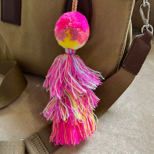 Bright pink pom pom tassel bag charm with layered multicolor tassels attached to a beige handbag