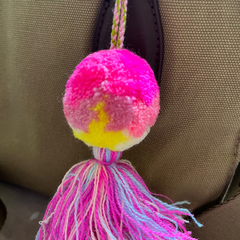 Hot pink pom pom tassel bag charm with layered multicolor tassels attached to a beige handbag