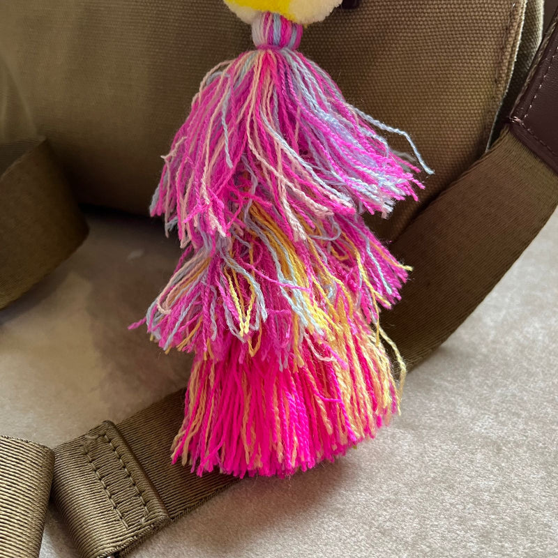 Hot pink pom pom tassel bag charm with layered multicolor tassels attached to a beige handbag