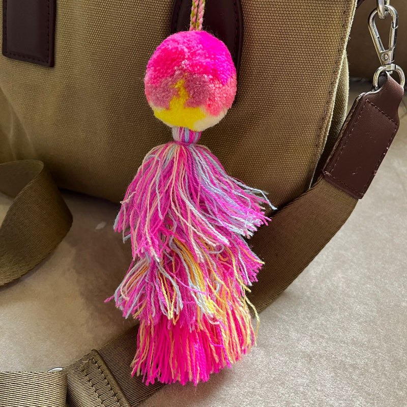 Hot pink pom pom tassel bag charm with layered multicolor tassels attached to a beige handbag