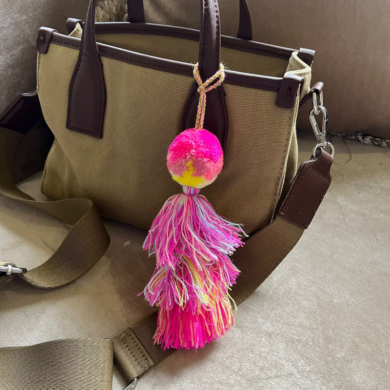 Hot pink pom pom tassel bag charm with layered multicolor tassels attached to a beige handbag