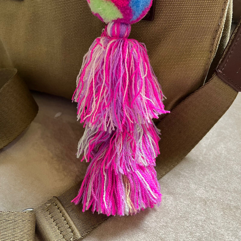 Rainbow pom pom tassel bag charm with layered pink tassels attached to a beige handbag
