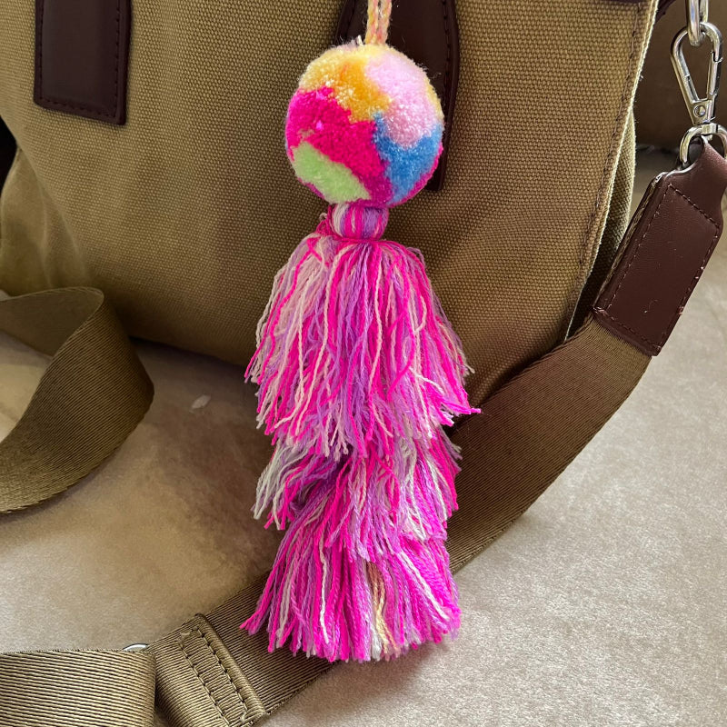 Rainbow pom pom tassel bag charm with layered pink tassels attached to a beige handbag