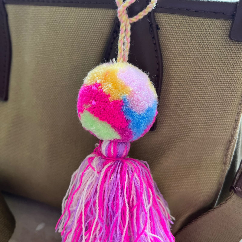 Rainbow pom pom tassel bag charm with layered pink tassels attached to a beige handbag