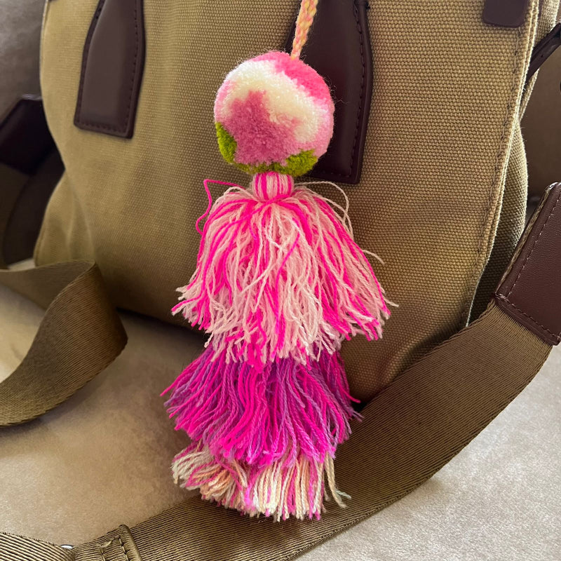 Blush pink pom pom tassel bag charm with layered pink tassels attached to a beige handbag