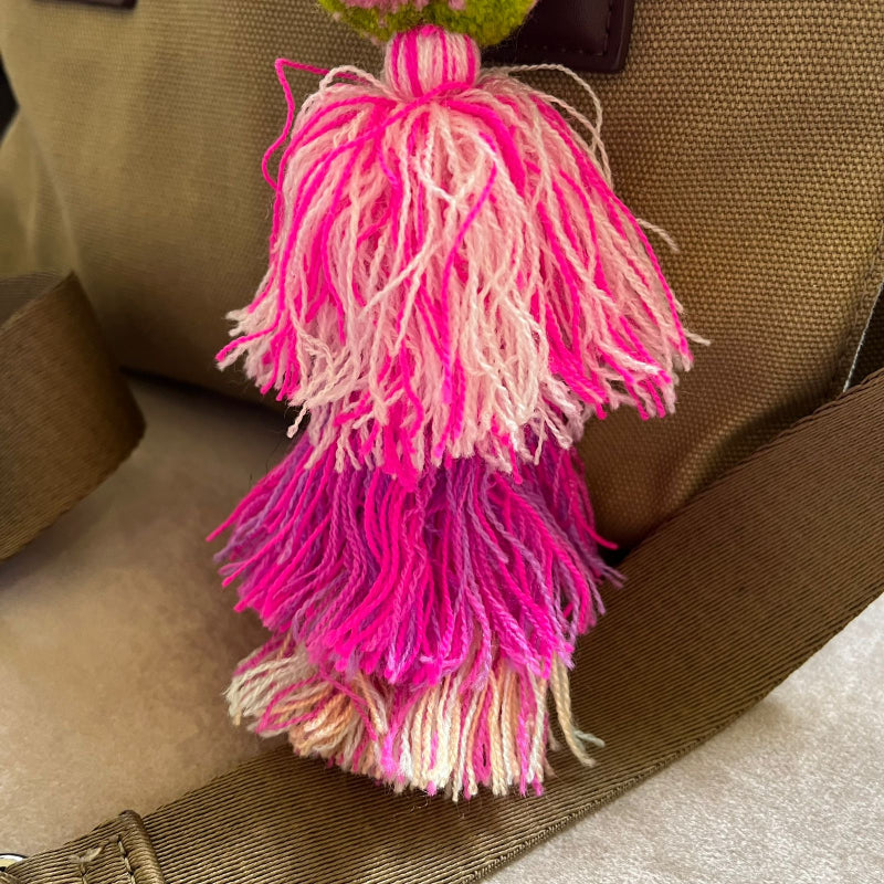 Blush pink pom pom tassel bag charm with layered pink tassels attached to a beige handbag