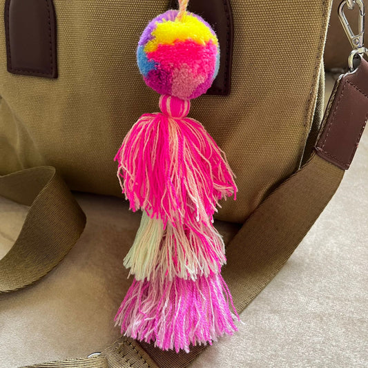 Neon pink pom pom tassel bag charm with layered multicolor tassels attached to a beige handbag