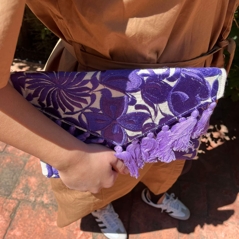 Purple floral embroidered clutch bag with tassels held by a person wearing a brown dress outdoors