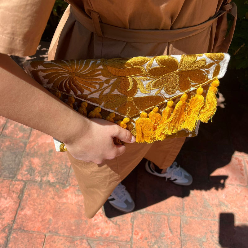 Mustard yellow floral embroidered clutch bag with tassels held by a person wearing a brown dress outdoors
