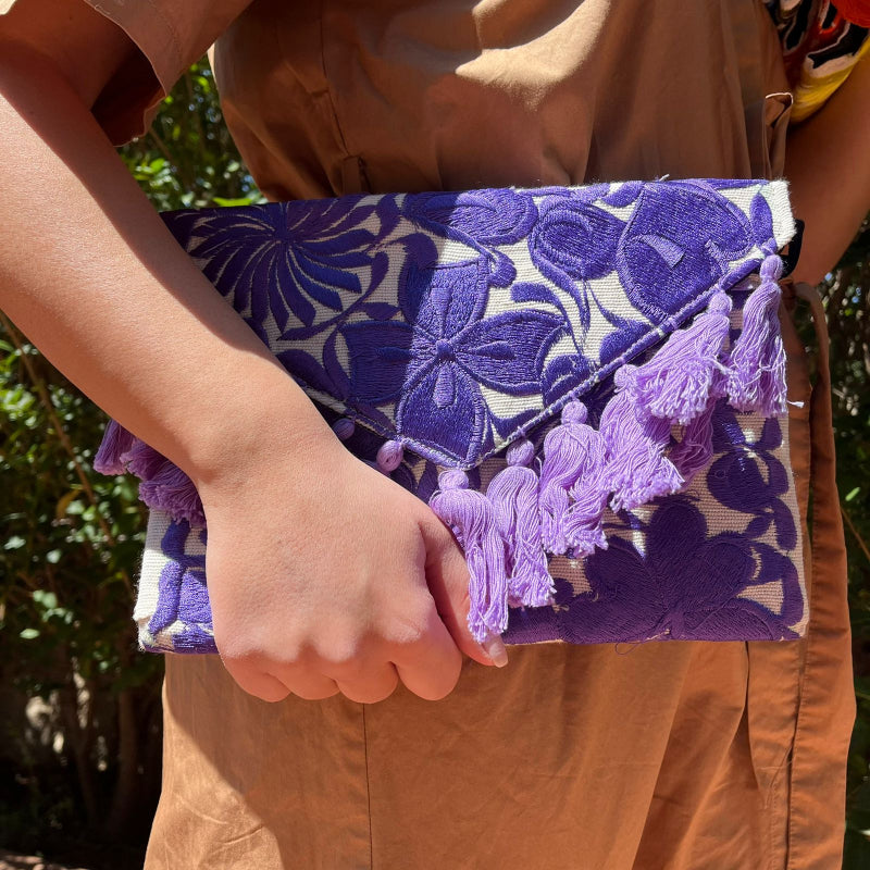 Purple floral embroidered clutch bag with tassels held by a person wearing a brown dress outdoors